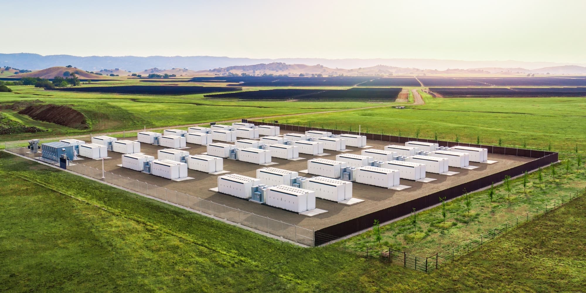 Vue aérienne d’un site de stockage par batteries : des rangées de conteneurs blancs sur une plateforme clôturée, au milieu de prairies