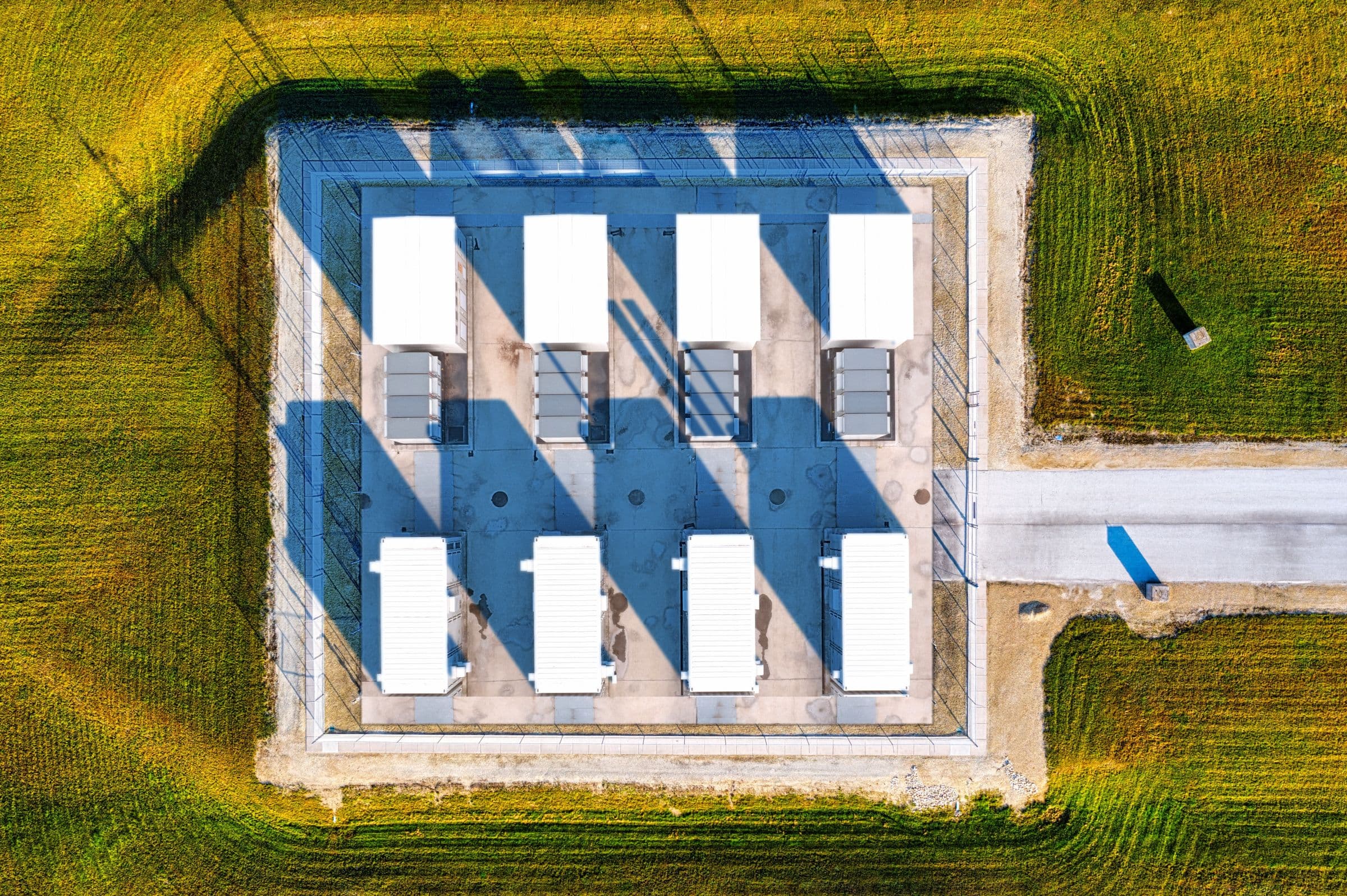 Vue aérienne d’un petit site de stockage par batteries : des conteneurs blancs sur une dalle clôturée, avec son chemin d’accès, au milieu d’une grande parcelle enherbée
